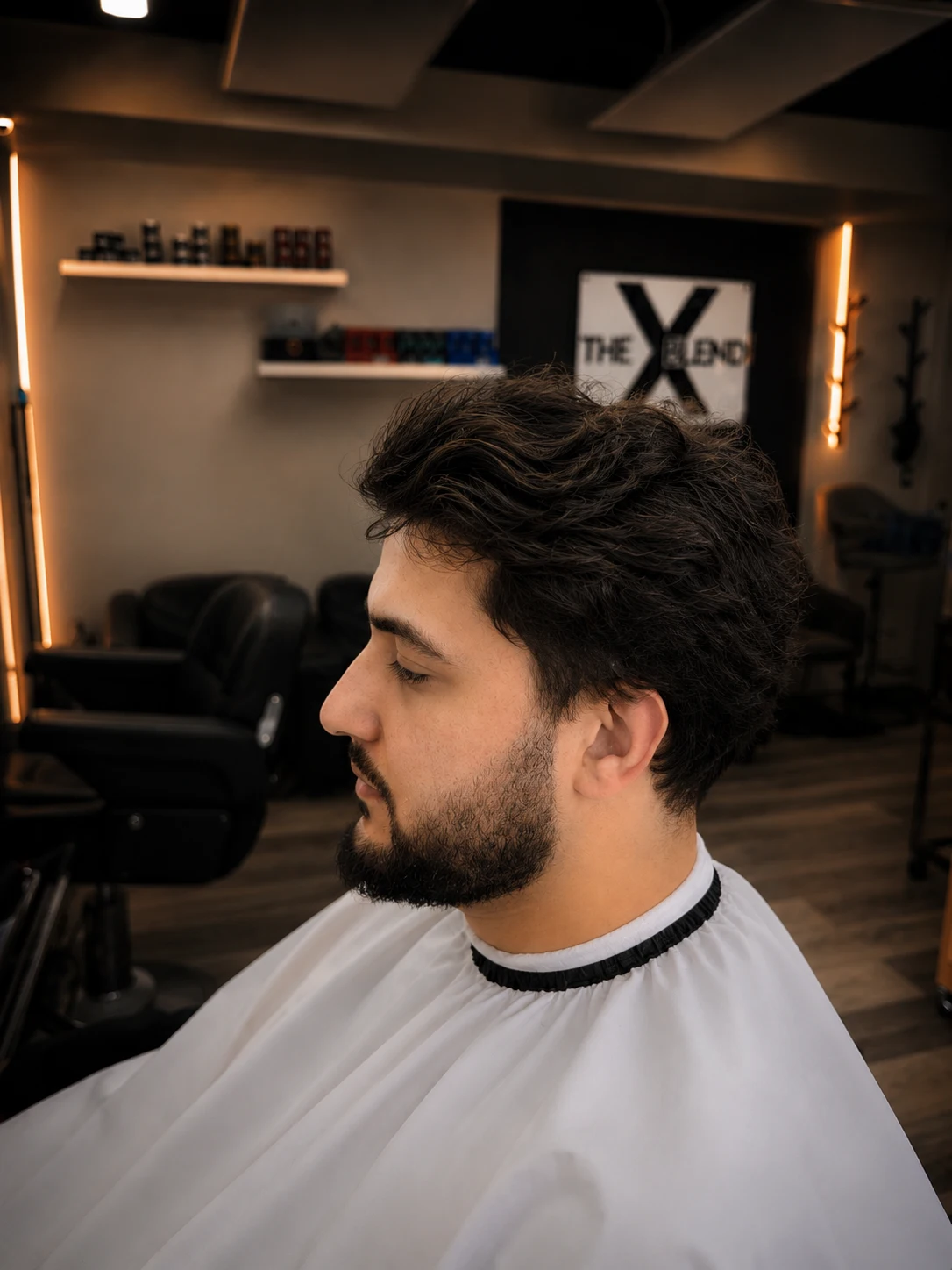 Side profile of a man with messy, thick dark hair and an untrimmed beard against a gray background.